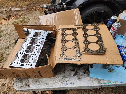 Automotive cylinder head and two head gaskets arranged on cardboard beside the vehicle.