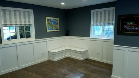 A finished basement corner with white wall paneling, an L-shaped bench beneath two windows, and dark upper walls.