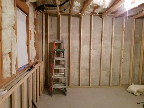 New wood wall framing along an unfinished basement corner, with insulation and a stepladder visible.