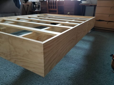 Uncovered platform-bed frame showing wood crosspieces, a recessed base, and plywood sides.