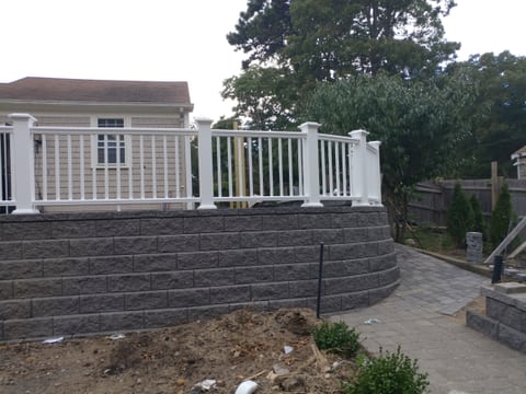 A curved wall of dark textured blocks with white railing above and a paved path along one side.