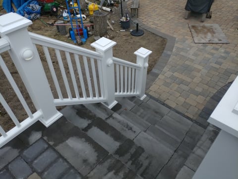 A view down gray paved steps between white railings to a lower paved area with contrasting border courses.