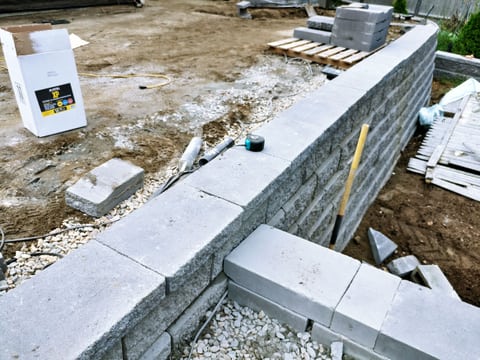 Close view of a block wall with cap pieces, a yellow level, and loose aggregate alongside the unfinished patio.